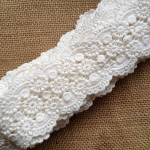 May include: White crochet lace trim with a delicate floral pattern. The lace is folded in half and has a scalloped edge.