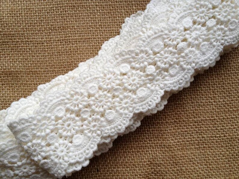 May include: White crochet lace trim with a delicate floral pattern. The lace is folded in half and has a scalloped edge.