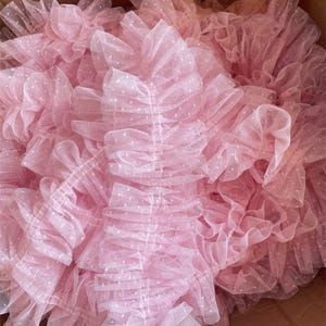 Puede incluir: Primer plano de un montón de tela de tul rosa con una textura delicada y con volantes. El tul está salpicado de pequeños lunares blancos, creando una apariencia suave y aireada. La tela está agrupada, mostrando su volumen y potencial creativo.