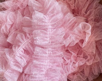 Può includere: Un bordo in tessuto di tulle rosa chiaro caratterizzato da un design denso a balze con un delicato motivo a pois bianchi. Il materiale trasparente e stratificato è raccolto in molteplici pieghe orizzontali, creando un aspetto voluminoso.