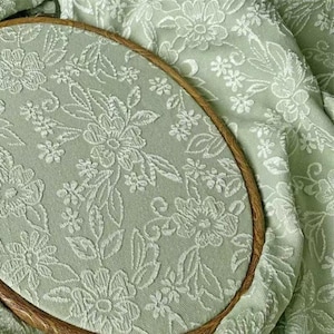May include: An oval embroidery hoop containing sage green fabric with a raised white floral pattern. The fabric features a dense, repeating design of various flowers and leaves. The wooden hoop frame is visible around the edge of the fabric.