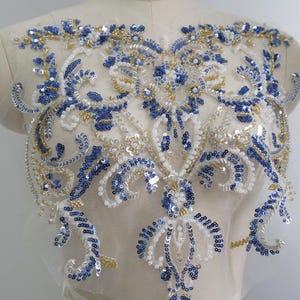 Op de afbeelding: Een decoratieve applicatie met ingewikkeld borduurwerk op transparante mesh-stof. Het ontwerp toont een symmetrisch bloemen- en krulpatroon, versierd met blauwe, witte en gouden pailletten, samen met doorzichtige en metallic kralen.