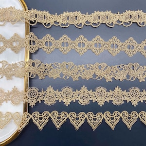 以下が含まれることがあります： 複雑なデザインの金色のレーストリムが5種類。トリムは暗い背景に一列に並んでいます。
