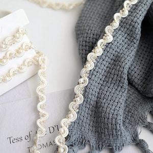 May include: White pearl and gold trim ribbon with a scalloped edge. The ribbon is shown on a gray fabric background.