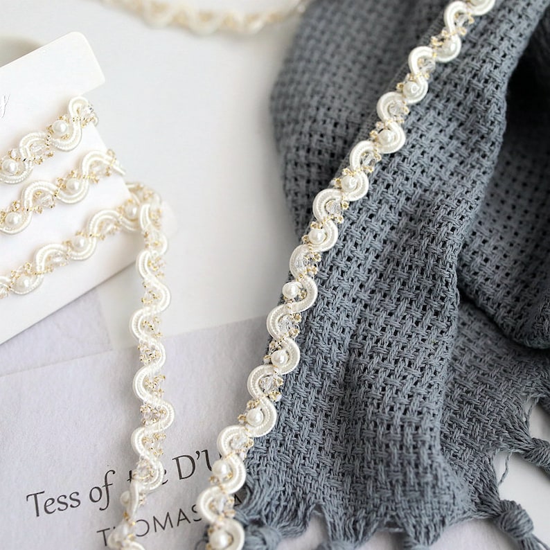 May include: White pearl and gold trim ribbon with a scalloped edge. The ribbon is shown on a gray fabric background.