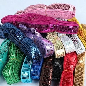 May include: A collection of multiple rolls of sequin trim in various colors including pink, magenta, dark blue, purple, green, teal, gold, silver, red, brown, and black. Each roll features a flat, flexible band covered in small, shiny sequins.