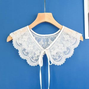 May include: A white lace collar with a scalloped edge and a tie closure. The collar is displayed on a wooden hanger against a blue background. The lace features a floral pattern.