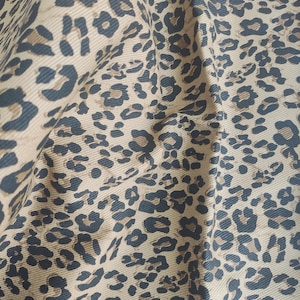 May include: Close-up of a tan fabric with a repeating leopard print pattern in dark brown and black. The fabric is slightly wrinkled, showing the texture and drape of the material. The print is detailed and covers the entire surface.