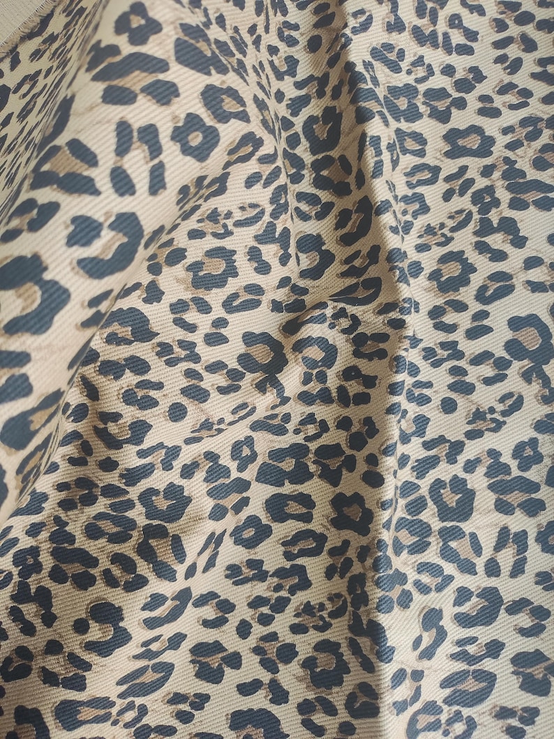 May include: Close-up of a tan fabric with a repeating leopard print pattern in dark brown and black. The fabric is slightly wrinkled, showing the texture and drape of the material. The print is detailed and covers the entire surface.