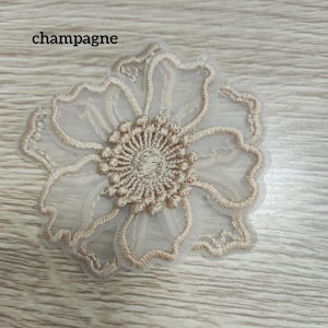 May include: A champagne-colored floral applique featuring intricate embroidery on a sheer mesh base. The design showcases a central circular cluster of stitches surrounded by five scalloped petals with defined edges. The text 'champagne' is visible.