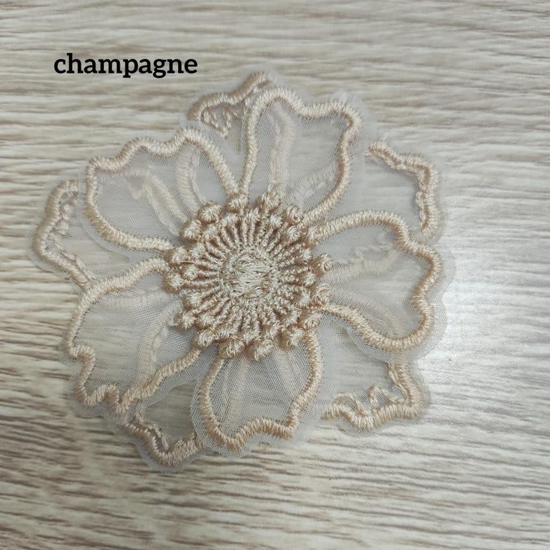 May include: A champagne-colored floral applique featuring intricate embroidery on a sheer mesh base. The design showcases a central circular cluster of stitches surrounded by five scalloped petals with defined edges. The text 'champagne' is visible.