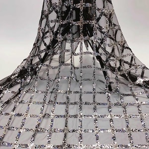 May include: Black sheer fabric with a grid pattern embellished with silver sequins. The fabric is draped, showcasing the intricate design and texture. The sequins catch the light, creating a shimmering effect, suitable for evening wear or decorative projects.