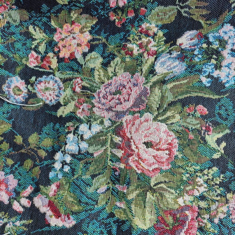 Puede incluir: Una tela tejida que presenta un denso patr&oacute;n floral sobre un fondo negro. El dise&ntilde;o incluye racimos de rosas rosadas, flores azul claro y varias hojas verdes, todo representado con un tejido de hilos texturizado y multicolor.