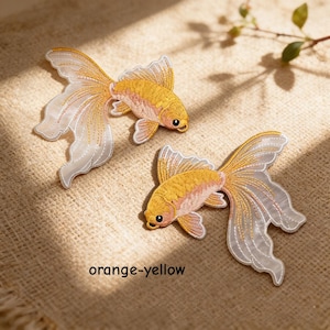 May include: Two embroidered goldfish patches featuring vibrant orange-yellow bodies and long, flowing, translucent white fins with delicate gold stitching. The text orange-yellow is printed below the fish. These decorative fabric appliques display intricate thread work and detailed scales.