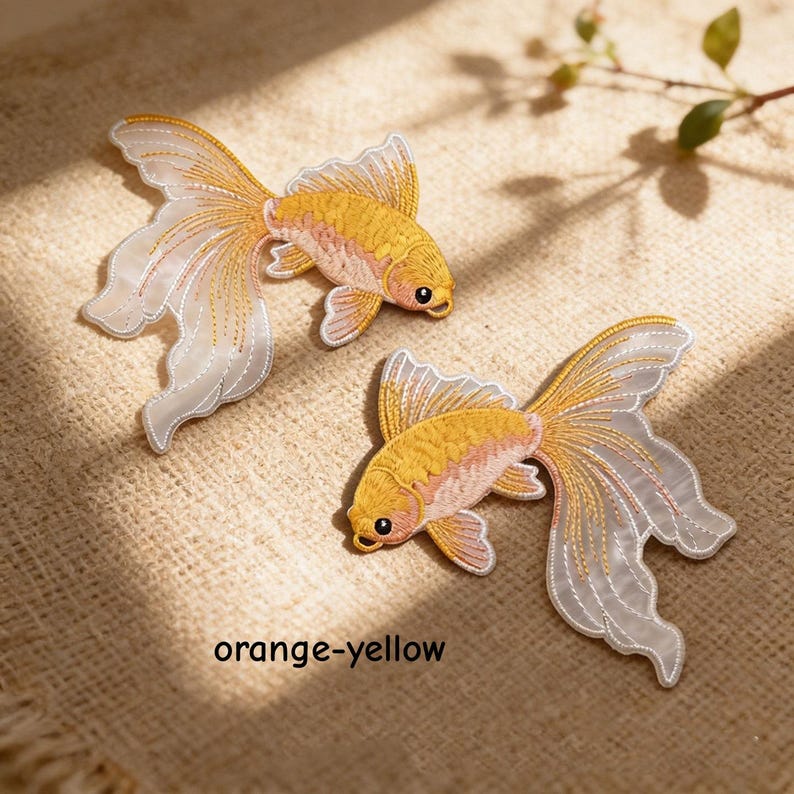 May include: Two embroidered goldfish patches featuring vibrant orange-yellow bodies and long, flowing, translucent white fins with delicate gold stitching. The text orange-yellow is printed below the fish. These decorative fabric appliques display intricate thread work and detailed scales.