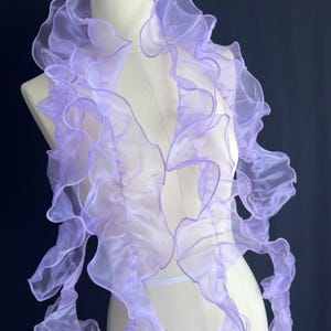 Puede incluir: Una bufanda de color morado claro hecha de tela de organza transparente y traslúcida. La bufanda presenta bordes voluminosos con volantes que crean una textura ondulada y en capas a lo largo del material, drapeada sobre un torso de maniquí blanco.