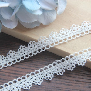 May include: White lace trim with a scalloped edge and delicate floral pattern. The trim is shown on a wooden surface.