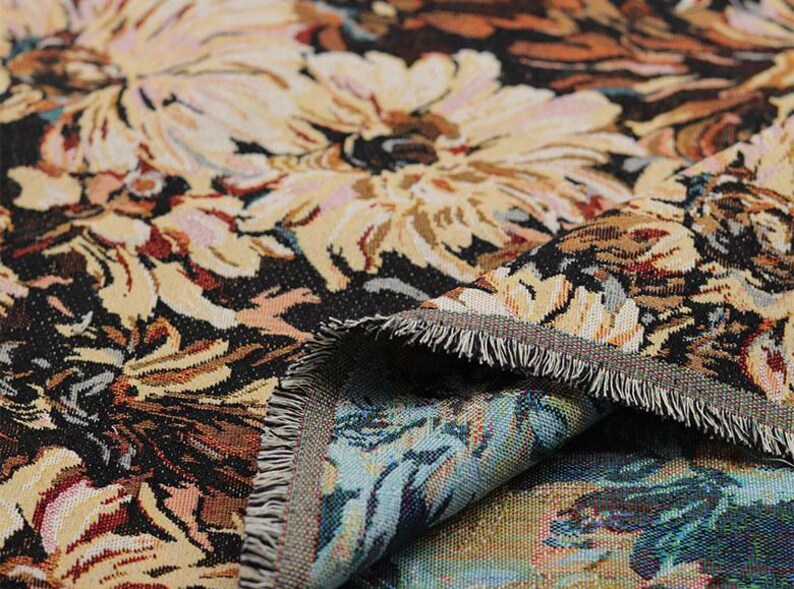May include: A woven tapestry fabric featuring a dense floral pattern with large cream and beige chrysanthemum-like flowers set against a dark black background. The fabric has a visible textured weave and a frayed edge revealing a blue underside.