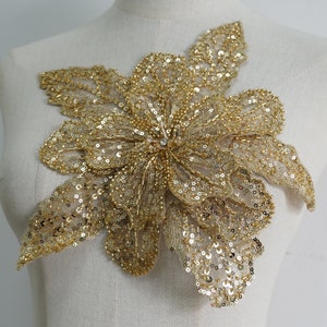 May include: A gold sequined flower applique with a delicate, layered design. The applique is perfect for adding a touch of sparkle and elegance to clothing, accessories, or home decor projects.