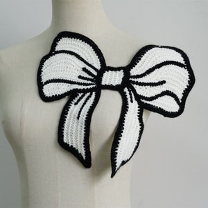 May include: White crocheted bow with black outlining. The bow is large and has a wide, flat shape.