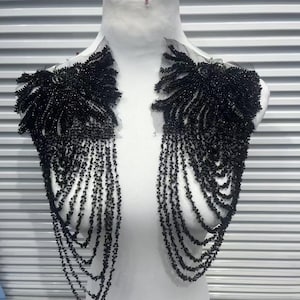 May include: Black beaded shoulder embellishments with a floral design. The embellishments feature a mesh base with intricate beadwork, creating a cascading fringe effect. The design is displayed on a white mannequin.