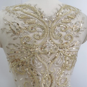 May include: Gold sequin and bead applique with an elaborate floral design. The applique has a symmetrical pattern with swirling details, set on a sheer, light-coloured fabric. The sequins and beads create a shimmering effect.