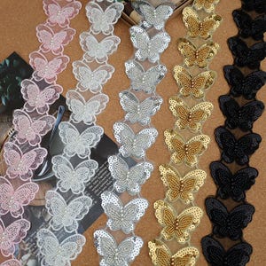 May include: Five vertical strips of butterfly-shaped lace trim arranged side by side. Each strip features a repeating pattern of butterflies embellished with small beads and sequins in pink, white, silver, gold, and black color variations.