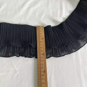 May include: Black pleated fabric trim with a scalloped edge, shown with a wooden ruler. The ruler measures up to 18 inches. The fabric is layered and has a delicate, sheer appearance. The trim is likely used for sewing or crafting projects.
