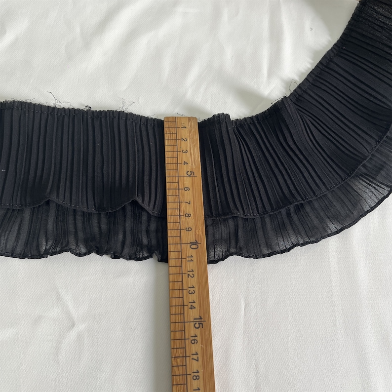 May include: Black pleated fabric trim with a scalloped edge, shown with a wooden ruler. The ruler measures up to 18 inches. The fabric is layered and has a delicate, sheer appearance. The trim is likely used for sewing or crafting projects.
