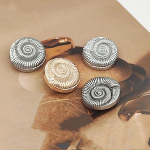 May include: Five metallic, fossil-shaped decorative objects. Four are silver-toned, and one is gold-toned. Each has a spiral design, resembling an ammonite shell. The objects are arranged on a light brown surface.