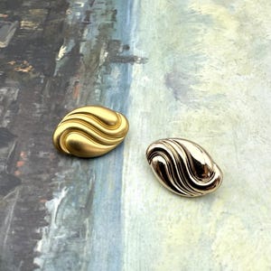 Puede incluir: Dos botones dorados de forma ovalada con un diseño ondulado y texturizado. Uno tiene un acabado mate, el otro es brillante. Estos botones son probablemente para ropa o accesorios.