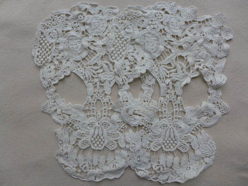 May include: Two white lace skull shapes, made of delicate floral patterns, are joined together at the top. The lace is intricate and detailed, with a delicate, almost translucent quality.