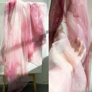 Puede incluir: Tela transparente en tonos rosa y blanco, drapeada sobre un caballete de madera. La tela tiene un efecto acuarela con áreas rosas más oscuras. La luz pasa a través de la tela, creando un aspecto suave y etéreo. La tela probablemente se utiliza con fines decorativos.