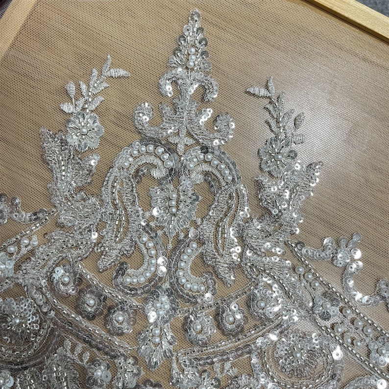 Pu&ograve; includere: Primo piano di un tessuto perlato argento e bianco con un motivo floreale. Il tessuto &egrave; fatto di paillettes e perle.
