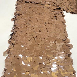 Puede incluir: Una tela de color marr&oacute;n caf&eacute; con una capa densa de lentejuelas circulares superpuestas. El material tiene un acabado met&aacute;lico reflectante y est&aacute; etiquetado con el texto Coffee Brown en la esquina superior izquierda, mostrando una textura uniforme.