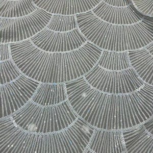 May include: Close-up of a fabric with a repeating pattern of fan-shaped designs. The fabric is a light gray color and is covered in small, round, silver sequins. The sequins catch the light, creating a shimmering effect.