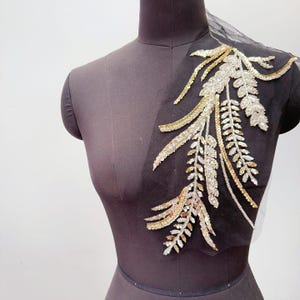 以下が含まれることがあります： 金と銀のスパンコールとビーズで刺繍された複雑な葉の模様が特徴の、黒いシースルーチュール生地のアップリケ。この装飾品は暗い色のマネキンに展示されており、詳細な植物のデザインとメタリックな仕上がりを際立たせています。