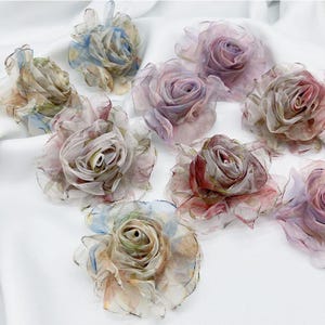 May include: A collection of artificial rose flowers crafted from translucent, layered fabric. Each flower features a delicate, multi-colored watercolor effect with soft hues of pink, blue, purple, and muted earth tones, arranged in a circular pattern.
