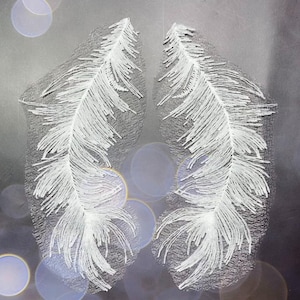 Peut inclure: Deux motifs blancs en forme de plumes, aux détails complexes, sur un fond gris dégradé avec des reflets circulaires flous. Les plumes ont un aspect délicat et texturé, suggérant une attention aux éléments naturels ou au design artistique.