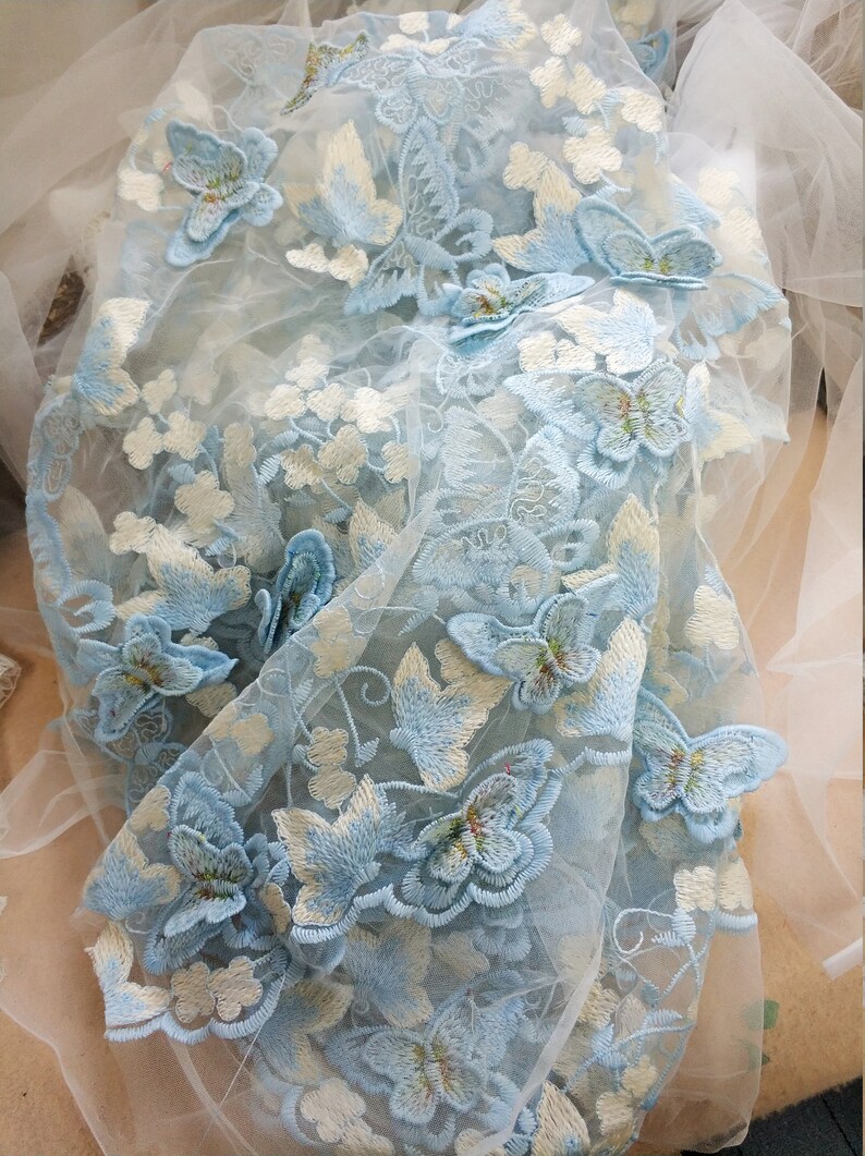 Pu&ograve; includere: Tessuto trasparente azzurro chiaro con farfalle ricamate. Le farfalle sono di diverse dimensioni e hanno un contorno bianco.