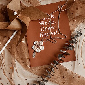 May include: A brown paper pinwheel with a white stick on a brown and white tulle fabric with gold stars. A brown card with the text "Think. Write. Draw. Repeat." and a gold necklace is on the fabric. A white flower with gold accents is on the card.