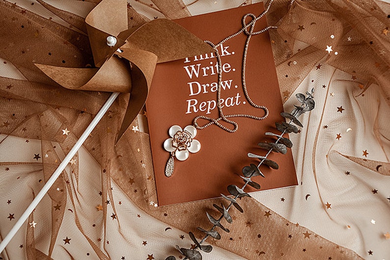 May include: A brown paper pinwheel with a white stick on a brown and white tulle fabric with gold stars. A brown card with the text "Think. Write. Draw. Repeat." and a gold necklace is on the fabric. A white flower with gold accents is on the card.