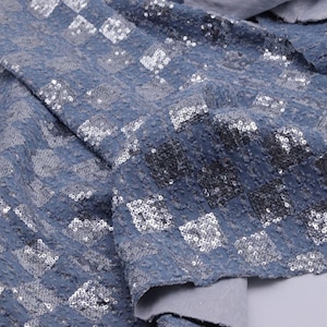 Puede incluir: Primer plano de una tela azul con un patrón de diamantes. La tela está cubierta de pequeñas lentejuelas plateadas que captan la luz. La textura parece suave y flexible, adecuada para prendas o proyectos decorativos.