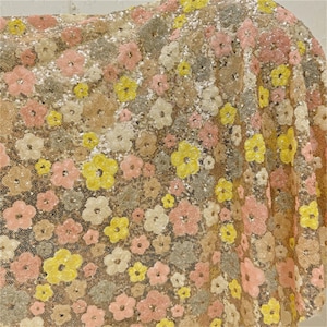 Peut inclure: Gros plan sur un tissu à motif floral. Le tissu est recouvert de paillettes et présente une palette de couleurs dorée, rose et jaune.