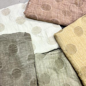 May include: Several folded fabric pieces in a palette of neutral tones, such as white, beige, and grey. Each piece is adorned with a large, circular pattern in a contrasting, muted shade. The fabric has a lightweight, textured appearance, possibly linen or cotton.