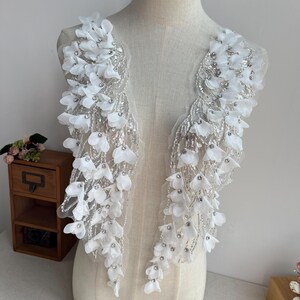 May include: Two white beaded floral appliques with silver sequins and rhinestones. The appliques are shaped like a cascade of flowers and are perfect for adding a touch of elegance to any garment.