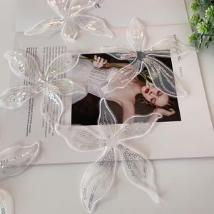May include: Several white, sequined flower appliques are scattered on a magazine page. The appliques have a delicate, translucent appearance with silver sequins. The background features a fashion magazine with a woman.