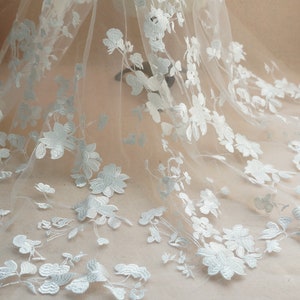 May include: White floral lace fabric with a delicate, light blue embroidery pattern. The fabric is sheer and has a soft, flowing texture.