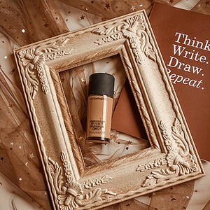 May include: A bottle of foundation in a light brown color with a black cap, inside a decorative, ornate, white frame. The frame is on a brown fabric with gold stars. A brown notebook with the text "Think. Write. Draw. Repeat." is visible in the background.