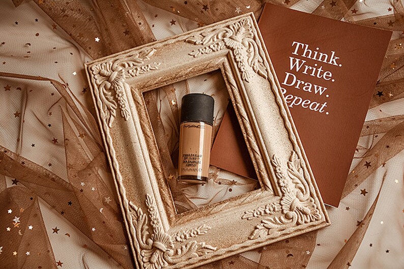 May include: A bottle of foundation in a light brown color with a black cap, inside a decorative, ornate, white frame. The frame is on a brown fabric with gold stars. A brown notebook with the text "Think. Write. Draw. Repeat." is visible in the background.
